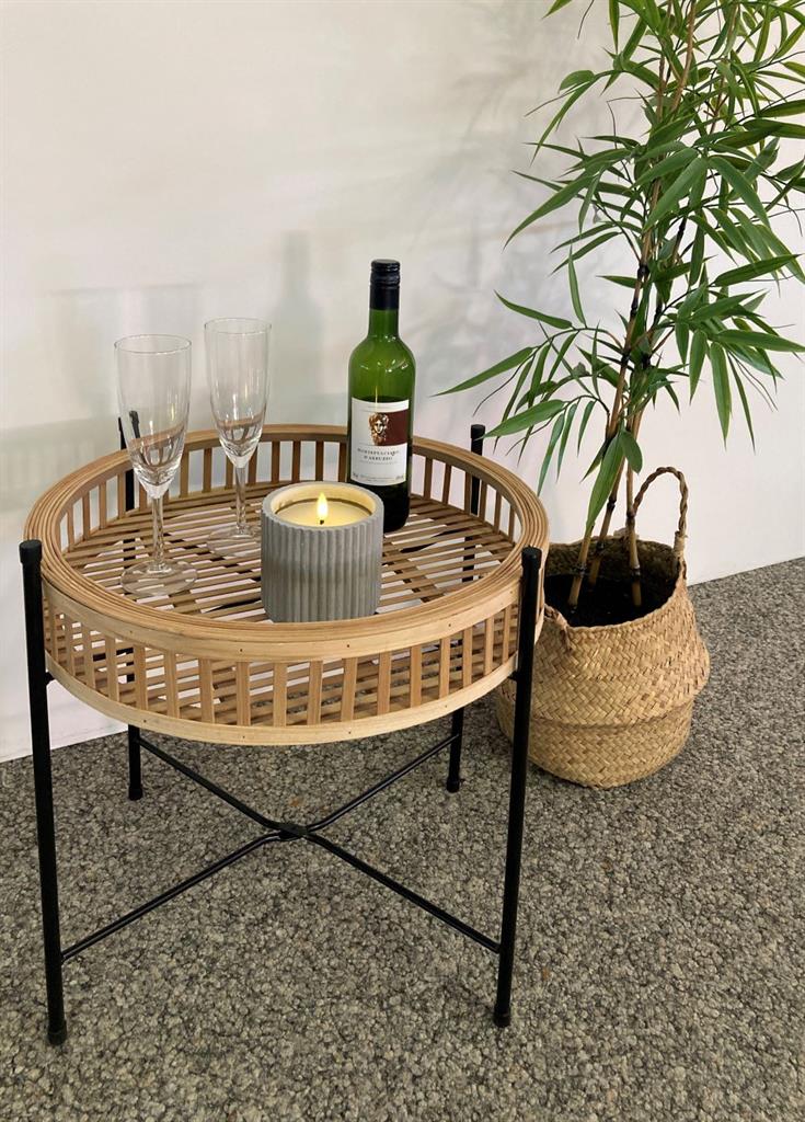 Runder Beistelltisch ZenTray aus Holz mit schwarzem Metallgestell, dekoriert mit Weinflasche, Gläsern und Kerze, daneben eine grüne Pflanze im Korb.